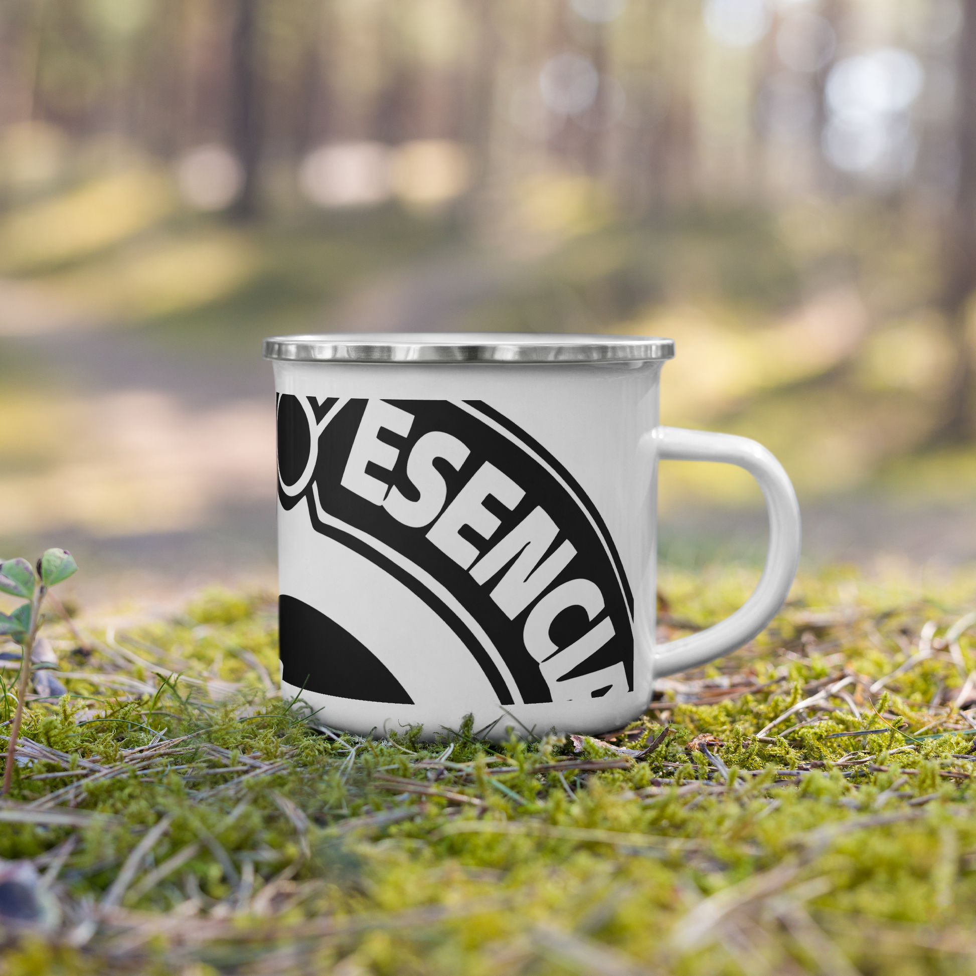 taza esmaltada de color blanco con el logo de la marca registrada tinta esencia impreso en negro. Sobre césped en un bosque